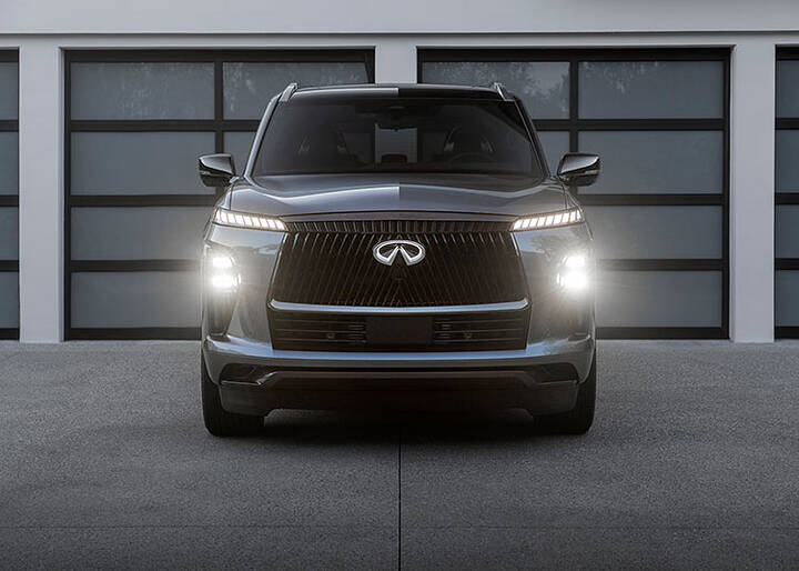 A front view of the 2025 INFINITI QX80 parked in front of multiple windows with the headlights and emblem lit.