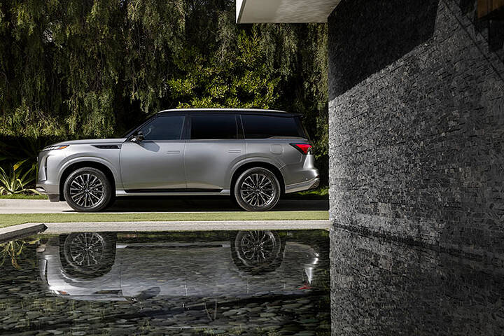A left side view of a silver 2025 INFINITI QX80 parked next to water and greenery, its reflection can be seen in the water.